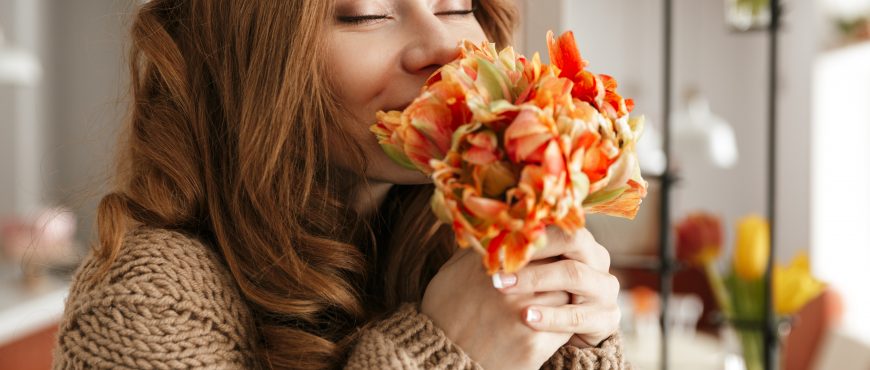 Photo of tender woman in sweater smelling beautiful flower with closed eyes while resting in restaurant or cafe